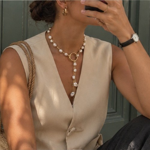 Mujer con un collar dorado, sentada frente a una puerta verde, con una taza en la mano.